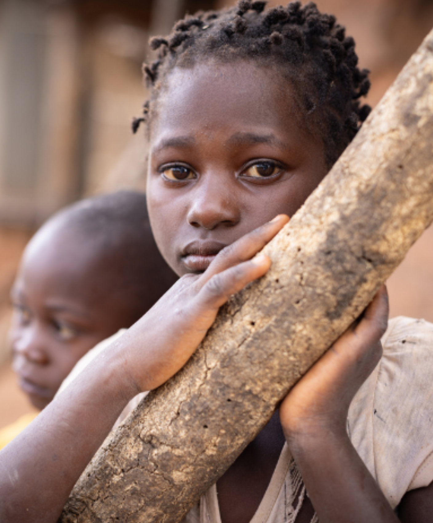 A young girl in Mozambique sits in front of her home, her face slightly hidden behind a wooden pole.