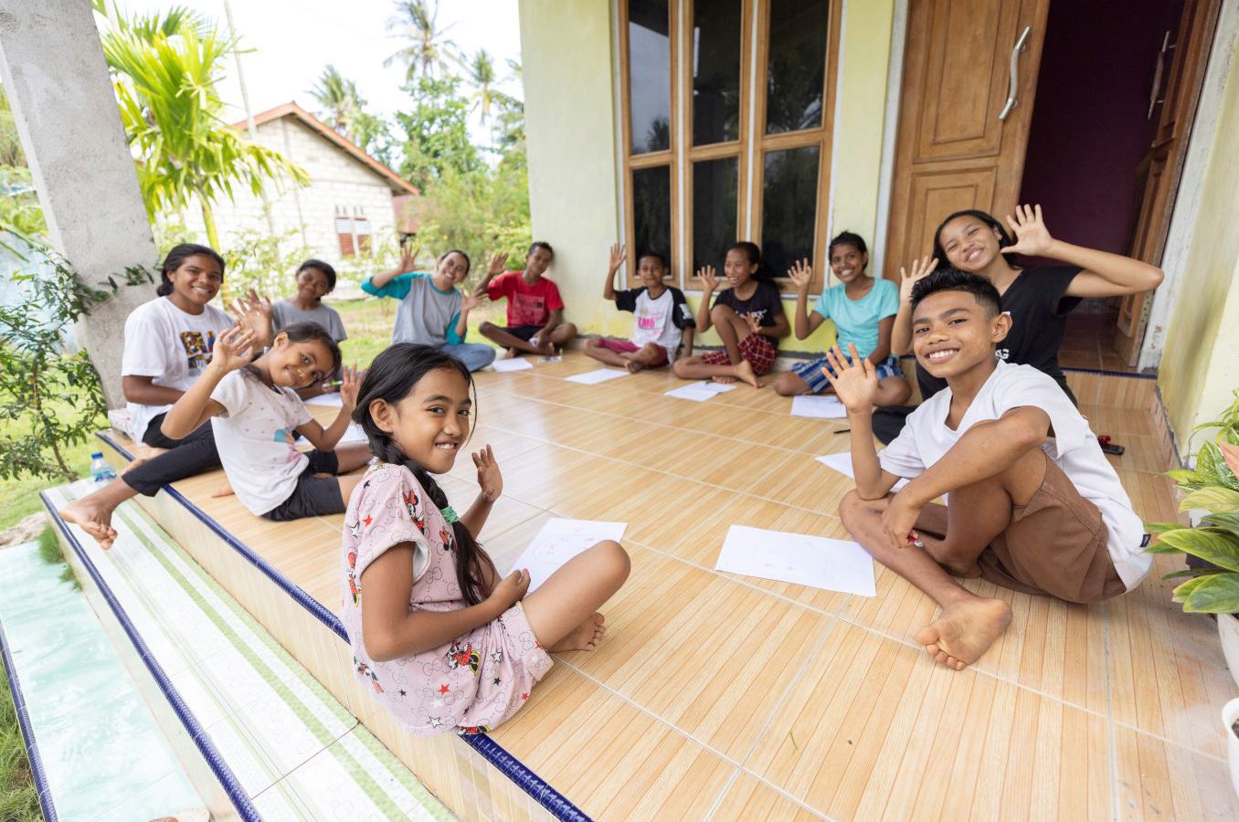 A group of children and youth sit in a circle on a wooden front porch in Indonesia, flashing peace signs and smiling at the camera.