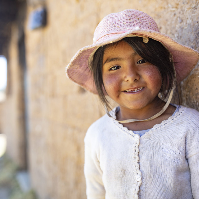 A young girl in Bolivia wearing a hat smiles in front of her home.