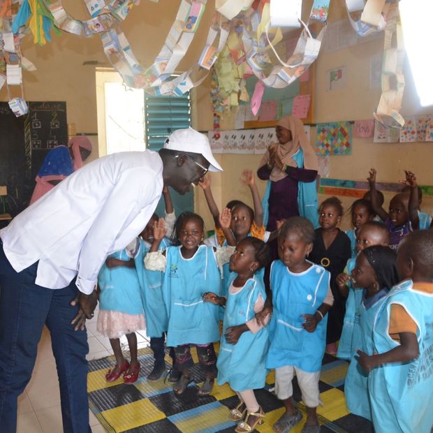 A man stands in a classroom in Senegal, leaning over a group of small children in school uniforms to talk to them. The children are all smiling up at him.